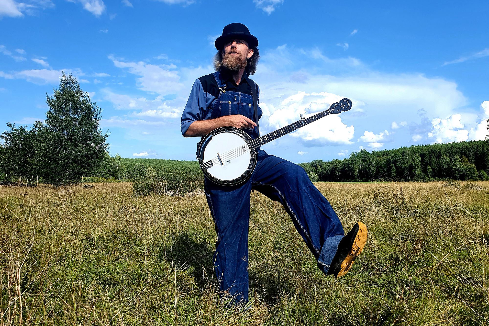En man i hatt som håller i en banjo, åker och blå himmel i bakgrunden.