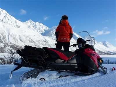 En person står ved en snøscooter i et snødekt fjellandskap med majestetiske fjell i bakgrunnen.