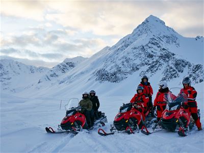 En gruppe gjester med snøscootere i et snødekt fjellandskap, omgitt av høye fjell.