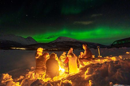 A group of guests gathered around a fire with the Northern Lights overhead.