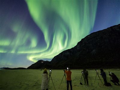Guests photograph a vivid green Northern Lights display at the foot of a mountain.