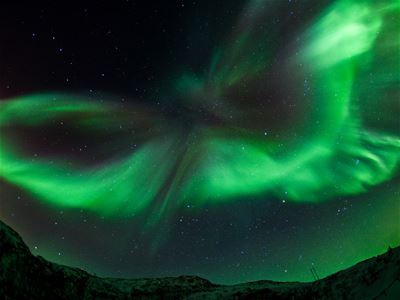 An intense green Northern Lights display moves across the dark Arctic sky above the mountains.