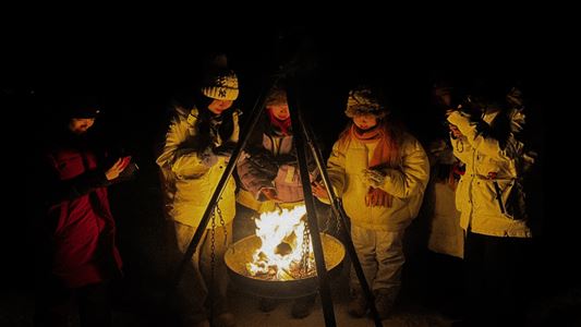 Gjester samles rundt bålet under en nordlysopplevelse i det arktiske vintermørket.Nordlys og fjell
