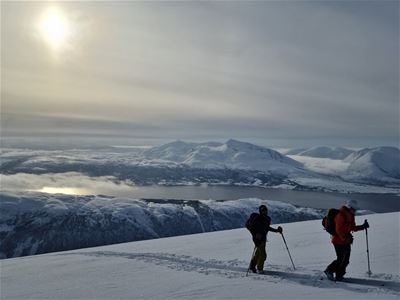 To skiløpere i et snødekt fjellandskap med utsikt over fjell og fjorder under en lys vinterhimmel.