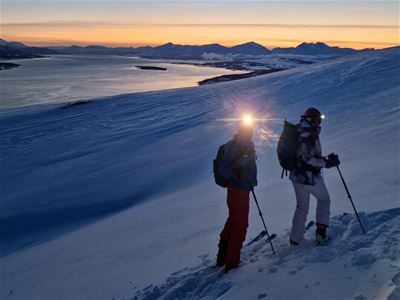 To skiløpere med ryggsekker og staver tar en pause i et åpent fjellandskap med snødekte fjell og lav vintersol.