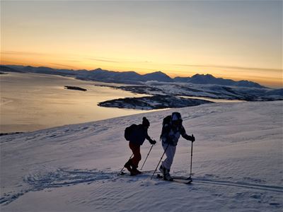 To skiløpere beveger seg oppover en snødekt fjellside med fjell og varmt kveldslys i bakgrunnen.