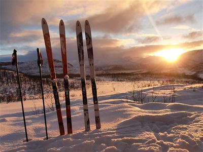 ki og skistaver står plantet i snøen med fjell og varmt kveldslys i bakgrunnen.