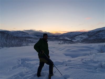 En person går på ski gjennom et snødekt landskap med fjell og dempet kveldslys i bakgrunnen.