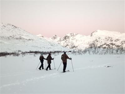 Tre personer går på ski gjennom et åpent, snødekt fjellandskap med fjell i bakgrunnen.