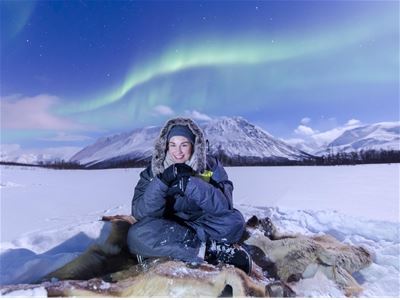 Gjest på reinsdyrskinn under nordlyset med snødekte fjell i bakgrunnen