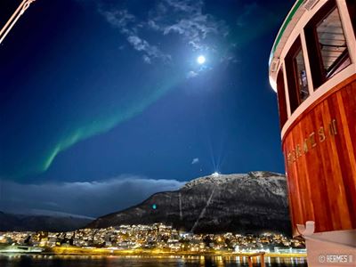 Nordlyset strekker seg over nattehimmelen ved Tromsøysundet, med måneskinn, fjell og lysene fra byen i bakgrunnen.