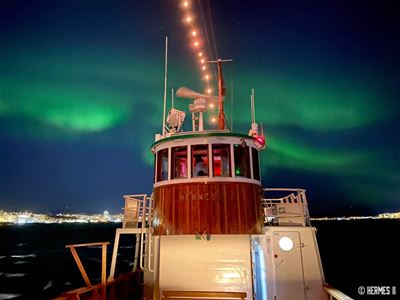 Den historiske trebåten Hermes II lyser opp i den arktiske natten, mens grønt nordlys danser på himmelen over Tromsø.