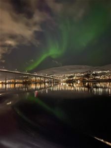 Grønt nordlys lyser opp nattehimmelen over Sandnessundbrua ved Tromsø, med refleksjoner i det rolige vannet.