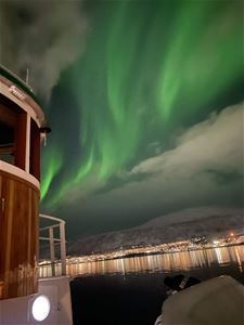 Kraftig grønt nordlys over Tromsø, sett fra dekket på Hermes II med lysene fra byen langs sjøen i bakgrunnen.