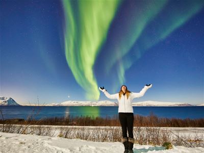 En gjest står under et sterkt, dansende nordlys i vinterlandskapet.