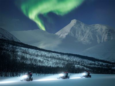 Guests drive snowmobiles past the mountains and the shining Northern Lights