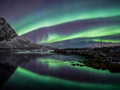 Green Northern Lights above a calm fjord and snow-covered mountains, with clear reflections of the aurora in the water.