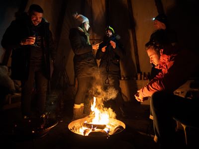Guests gathered around a warm campfire during a Northern Lights tour, wearing winter clothing and enjoying hot drinks.