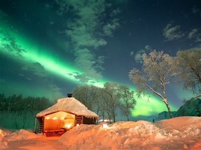 En opplyst hytte i et snødekt vinterlandskap med grønt nordlys over himmelen og snødekte trær rundt campen.