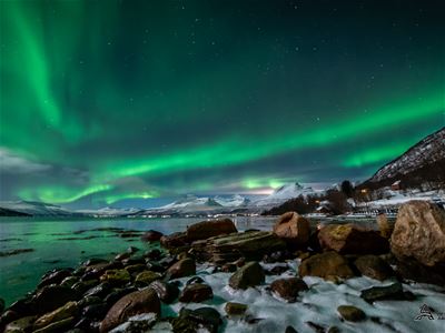 Green Northern Lights stretching across an Arctic coastal landscape with mountains, rocks and the sea.