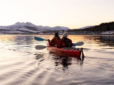Gjester padler kajakk på rolig vann med fjellene rundt Sommarøy i bakgrunnen.