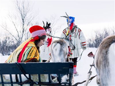 Sámi host in traditional clothing sitting on a reindeer sled