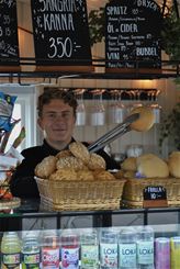 En leende person bakom en bageridisk med korgar av frallor och kritskyltar med dryckesmeny ovanför.