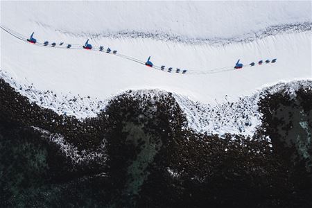 Dog sled teams travelling through a snow-covered Arctic landscape, seen from above with dark mountain formations in the foreground.
Dog sledding