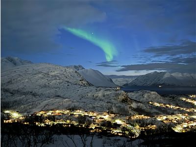 The Northern Lights illuminate the sky above snow-covered mountains, a fjord and a small coastal community.