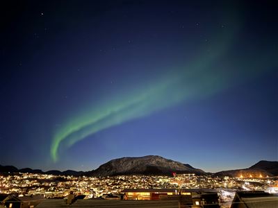 Green Northern Lights stretch across the sky above an illuminated coastal community and mountain.