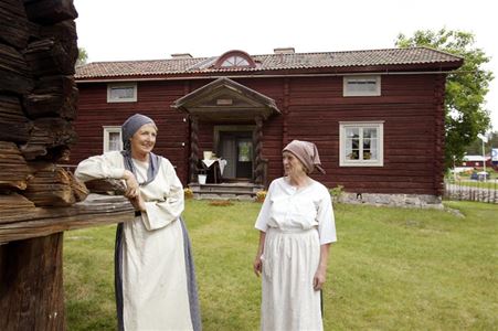 Two women in older clothes in front of a red timber bilding in two floors.