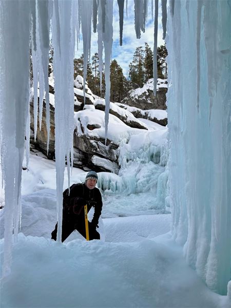 Person står mellom høye istapper ved en frossen foss i et snødekt skogsområde.