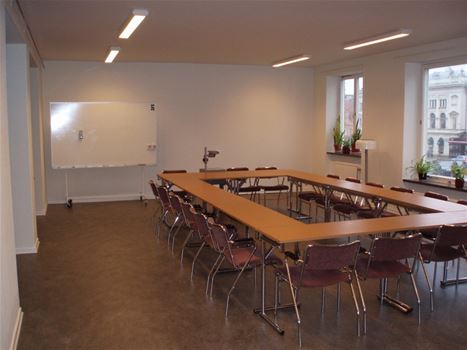 Ett vitt konferens rum med ett rektangel bord med stolar och en whiteboard tavla