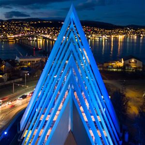 Ishavskatedralen opplyst på kveldstid med Tromsø og fjellene i bakgrunnen.