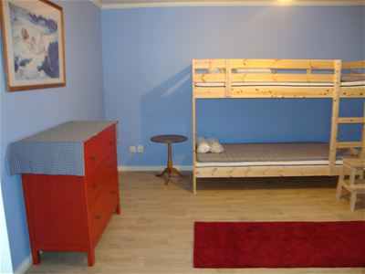 Blått rum med våningssäng av furu, röd byrå och röd matta på golvet.