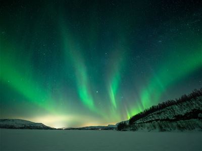 Vibrant green Northern Lights dance across the Arctic night sky above a snowy landscape. The guided Northern Lights chase with Tromsø Arctic Reindeer offers guests the opportunity to witness this natural phenomenon.