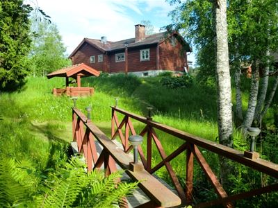 Cabin with a green lawn, a  water well and a red painted bridge railing. 