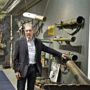 A man in a dark jacket and white shirt stands and watches an exhibition of different weapons.