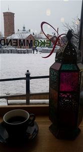 Utsikt från ett fönster, snö på backen utanför, i fönstret en svart kopp med svart fat brevid den en lykta i svart mide och glas i olika färger.