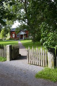 An open gate, a gravel walk, garden, a red timber building in two floors.