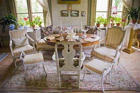 Ett ljust rum med en soffa, ett runt bord, tre vita stolar, två fönster med pelagonier.