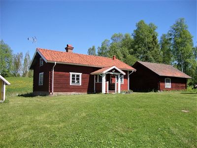 Gårdsplan med grön gräsmatta, rött timmerhus med vita knutar och en mindre stuga bredvid. 
