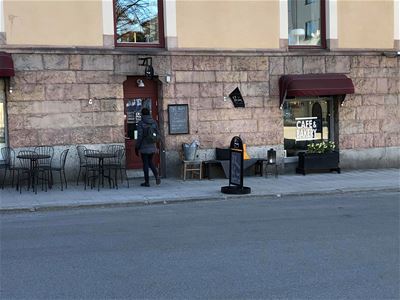 Exteriörbild, svarta stolar och bord som står på trottoar, svart stående skylt, vit text på ett fönster café o bageri.
