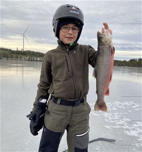 Boy with a fish in his hand standing on the ice wearing a helmet.