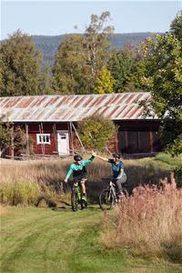Två cyklister gör en high five. I bakgrunden syns en gärdesgård och en röd ladulänga.