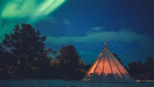 Opplyst lavvo i vinterlandskap under grønt nordlys.
