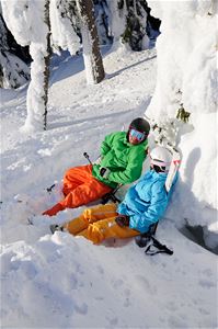 Två skidklädda personer sitter i snön och vilar, solar.